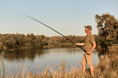 Full length photo of elderly man wearing casual attire and hat holding fishing rod in hands, looking at bobber, enjoying rest and silence.