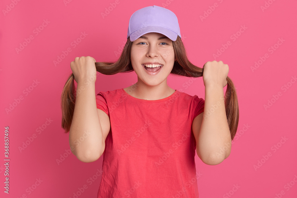 Young woman dresses red t shirt and baseball cap posing isolated over pink background, holding her hair in hands like wants make ponytails, looks excited directly at camera. Young woman dresses red t shirt and baseball cap posing isolated over pink background, holding her hair in hands like wants make ponytails, looks excited directly at camera.