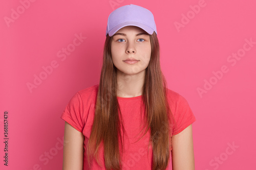 Beautiful woman wearing baseball cap and t shirt posing isolated over pink background, looks at camera with serious expression on face, having long straight hair.