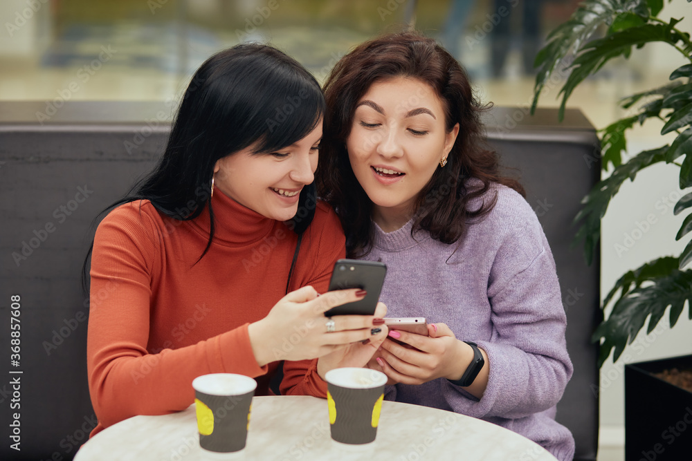 Two excited young girls using mobile phones while sitting at table in cafe and drinking hot beverages, brunette girls looking at smart phone screen and reading something interesting. Two excited young girls using mobile phones while sitting at table in cafe and drinking hot beverages, brunette girls looking at smart phone screen and reading something interesting.