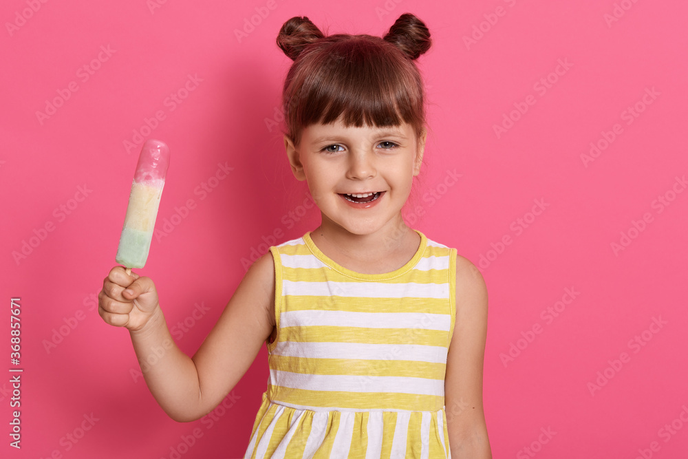 Smiling little girl with ice cream posing isolated over rose background, looking at camera with joyful expression, holding water ice in hands, funny female kid with knots, wearing summer dress. Smiling little girl with ice cream posing isolated over rose background, looking at camera with joyful expression, holding water ice in hands, funny female kid with knots, wearing summer dress.