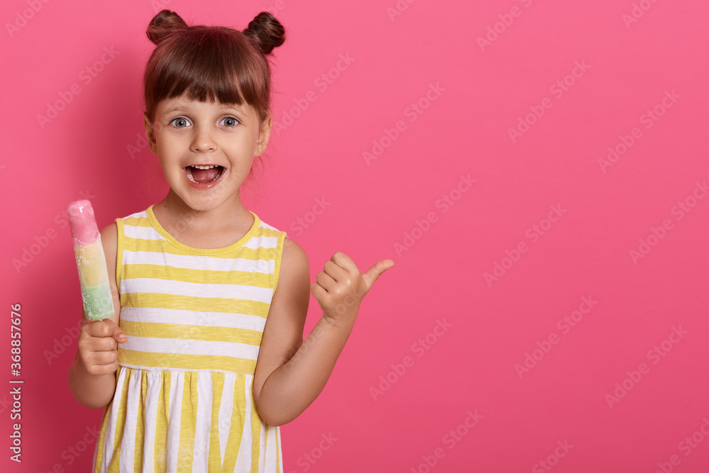 Happy excited little kid with ice cream looking at camera with widely opened mouth, pointing thumb aside, funny girl with knots looks at camera, copy space for advertisement. Happy excited little kid with ice cream looking at camera with widely opened mouth, pointing thumb aside, funny girl with knots looks at camera, copy space for advertisement.