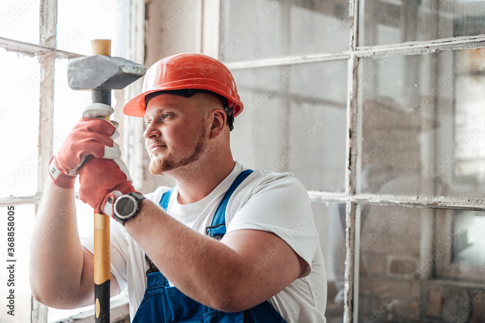 Worker, looking out the window, stands at the construction site. He is wearing a red safety helmet and he holds a sledgehammer for repairs in his hands. Worker, looking out the window, stands at the construction site. He is wearing a red safety helmet and he holds a sledgehammer for repairs in his hands.