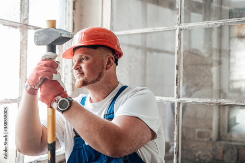 Worker, looking out the window, stands at the construction site. He is wearing a red safety helmet and he holds a sledgehammer for repairs in his hands.