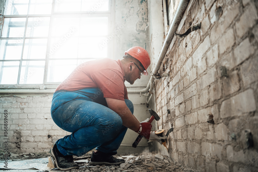 Professional worker in a safety helmet strikes old plaster from brick wall Professional worker in a safety helmet strikes old plaster from brick wall