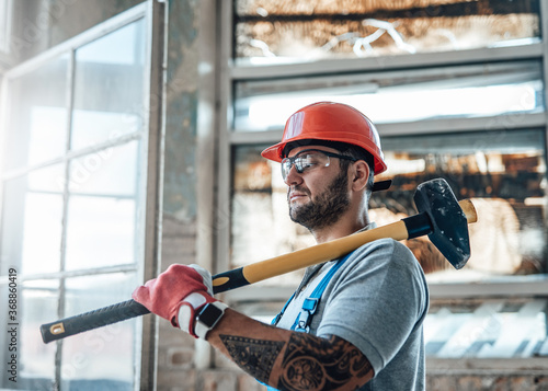 Attractive constructional worker with tattos stands near the window at a construction site. He is wearing a red safety helmet and he holds a sledgehammer for repairs in his hands.