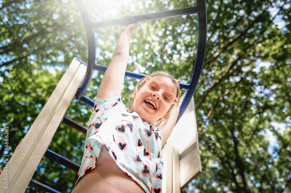 Obraz premium Happy teenage girl playing on the playground. Child girl hanging upside down on the playground. Smiling teenager girl playing at a sports construction
