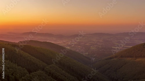 Timelapse of the sunset on the hillside and mountainous landscape below which lie several smaller villages the sun changes color from orange to dark blue during the west of the Beskydy region.