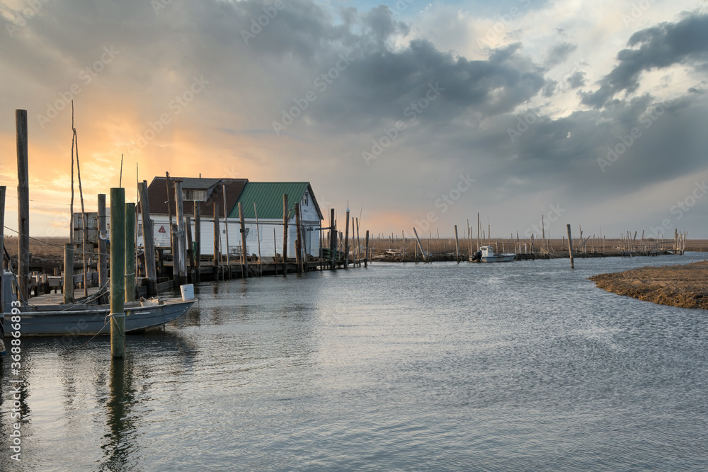 Nautical scene with a bait house, pilings, a dock and a small boat at sunset.