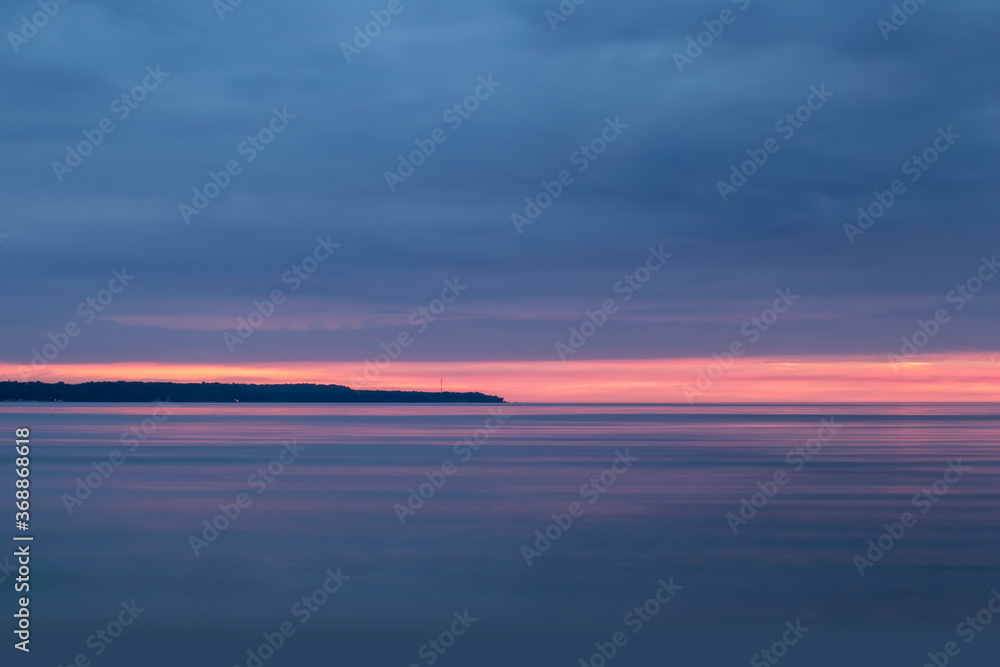 A beautiful pink and blue sunset over the sea and an island or a peninsula with a mobile tower visible between trees, copy space in the cloudy sky