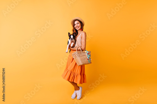 Full-length portrait of romantic young lady with summer hat holding dog. Stunning red-haired girl in long skirt posing with french bulldog.