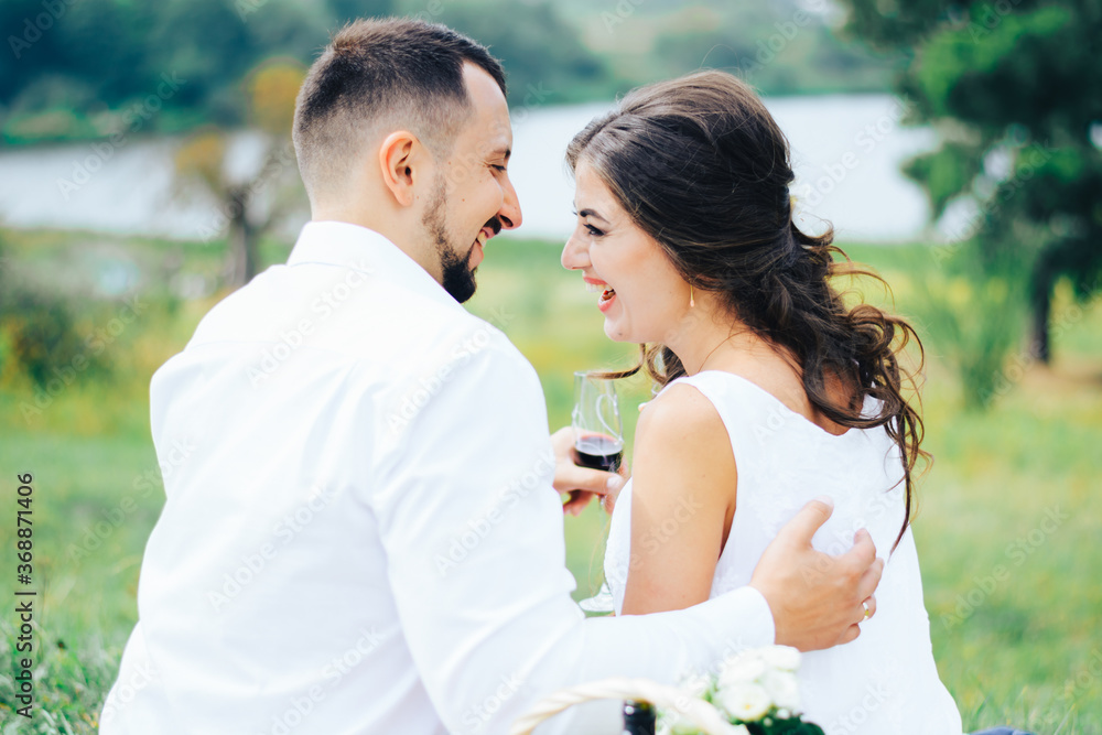 Beautiful couple drinking wine at a picnic by the lake. Honeymoon concept. Rear view. Beautiful couple drinking wine at a picnic by the lake. Honeymoon concept. Rear view.