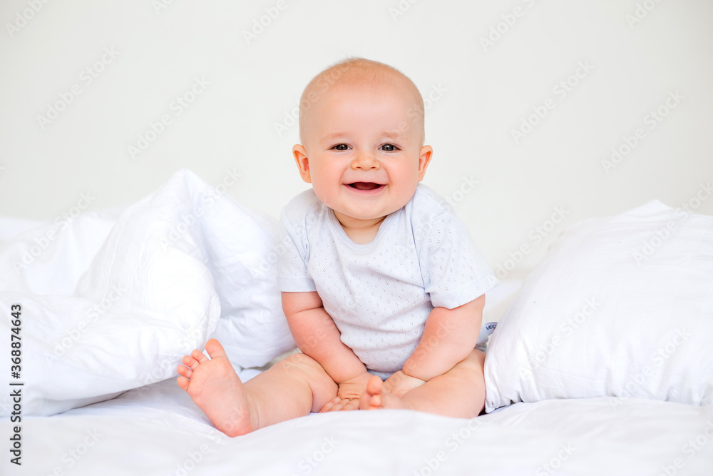 Cute baby boy 6 months smiling in a white bodysuit sitting on a bed on white bedding Cute baby boy 6 months smiling in a white bodysuit sitting on a bed on white bedding