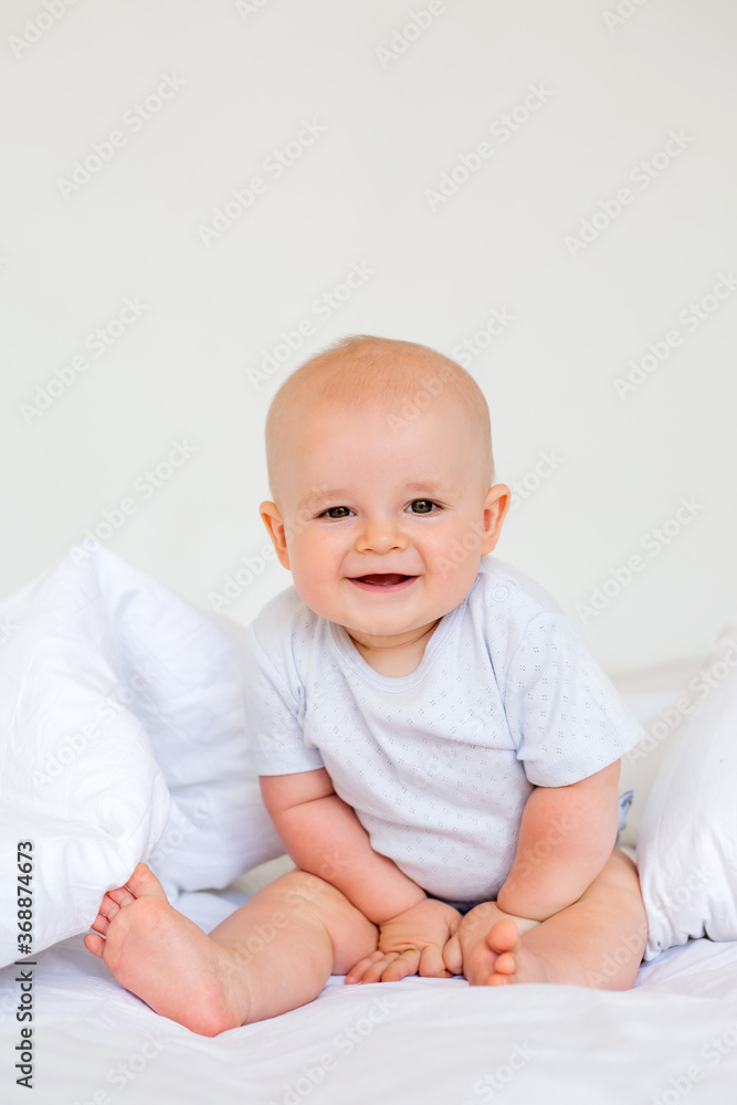 Cute baby boy 6 months smiling in a white bodysuit sitting on a bed on white bedding Cute baby boy 6 months smiling in a white bodysuit sitting on a bed on white bedding