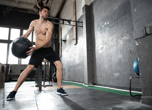 Young strong sweaty focused fit muscular man doing throwing medicine ball up on the wall for crossfit training hard core workout in the gym