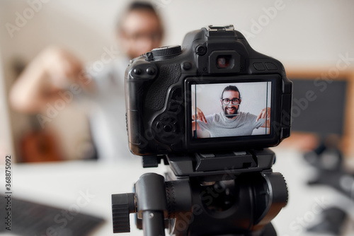 Young male blogger pointing down, asking followers to subcribe while recording video blog or vlog at home studio. Focus on camera screen