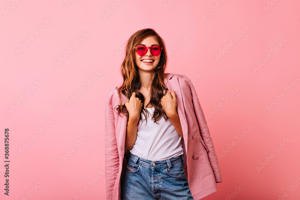 Lovable ginger girl in elegant pink jacket laughing in studio. Winsome young woman in trendy accessories standing on rosy background. Lovable ginger girl in elegant pink jacket laughing in studio. Winsome young woman in trendy accessories standing on rosy background.