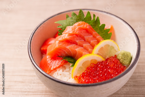 raw pink salmon sashimi and red salmon roe ikura over white rice with lemon wedges in a ceramic bowl