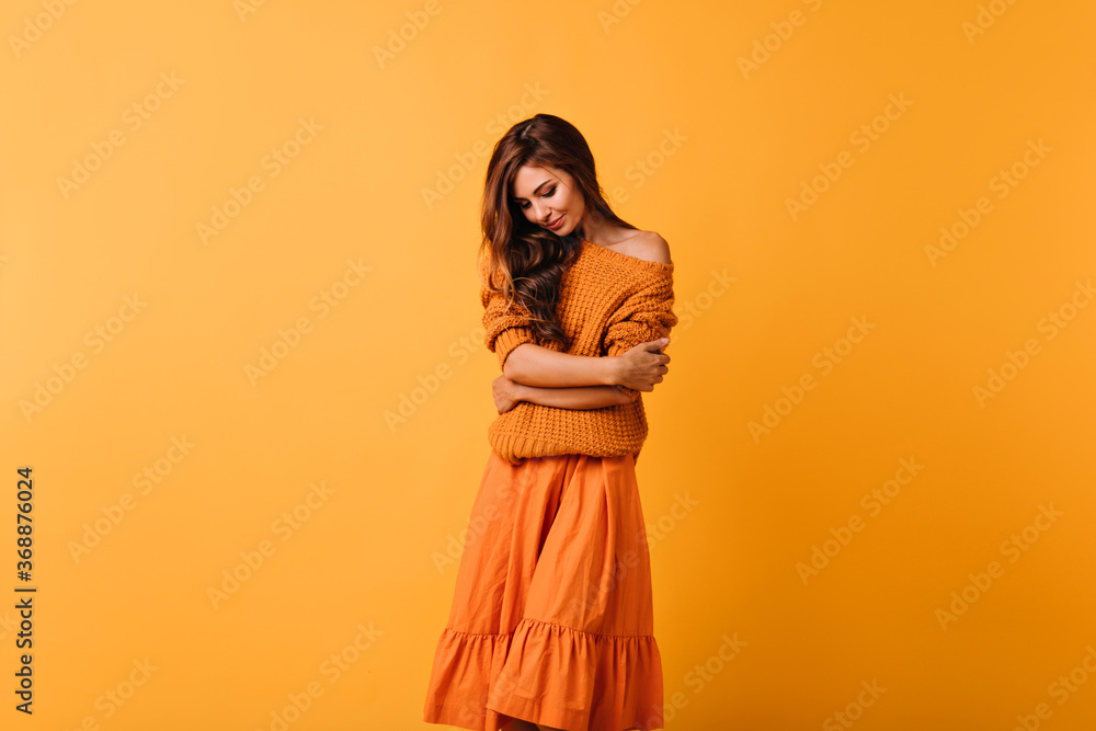 Romantic dark-haired lady in cozy sweater posing on yellow background. Good-humoured female model in orange skirt looking down.