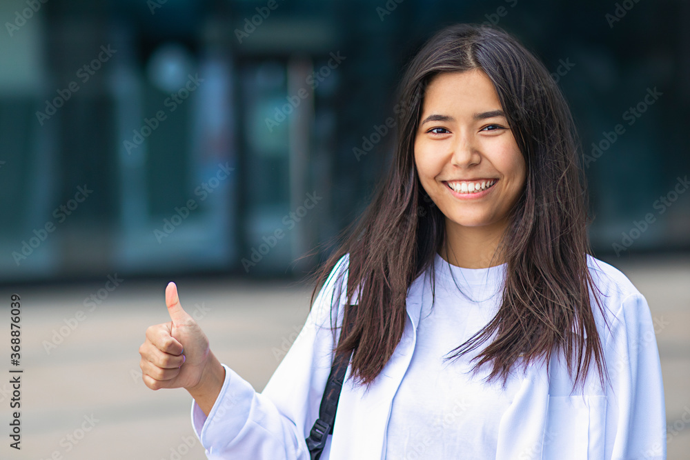 Female Asian ethnic businesswoman laughing, smiling wide with white teeth and showing a finger gesture, cool sign, thumb up, like. Happy student girl in white shirt outdoors. Business, approval Female Asian ethnic businesswoman laughing, smiling wide with white teeth and showing a finger gesture, cool sign, thumb up, like. Happy student girl in white shirt outdoors. Business, approval