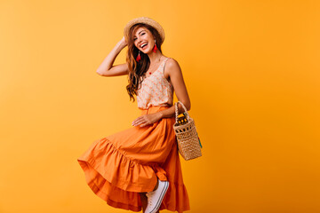 Joyful european lady in summer hat dancing on yellow background. Debonair girl in long skirt laughing while posing in studio.