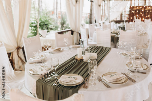 Wedding table setting. Banquet table with white tablecloths served with white plates, silver cutlery, white napkins and wine glasses. olive colore drapery on a white set table © Tetiana
