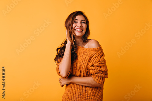 Adorable woman in orange attire touching her brown wavy hair. Laughing blithesome girl posing on yellow background.