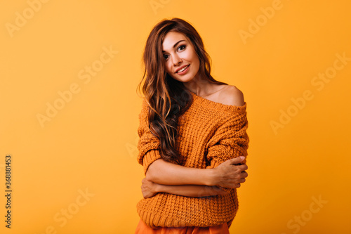 Interested long-haired lady in orange sweater looking to camera. Debonair european female model standing on yellow background with arms crossed.