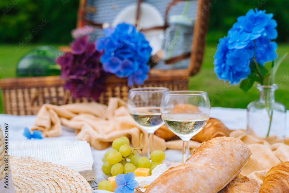 Summer picnic on sunny day with bread, fruit, bouquet hydrangea flowers, glasses wine, straw hat, book and ukulele. Picnic basket on grass with food and drink on white knit blanket. Selective focus
