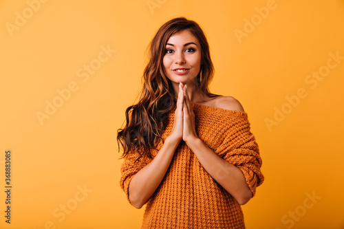 Dreamy young lady in stylish orange sweater posing with cute smile. Indoor photo of adorable caucasian girl with long wavy hair.