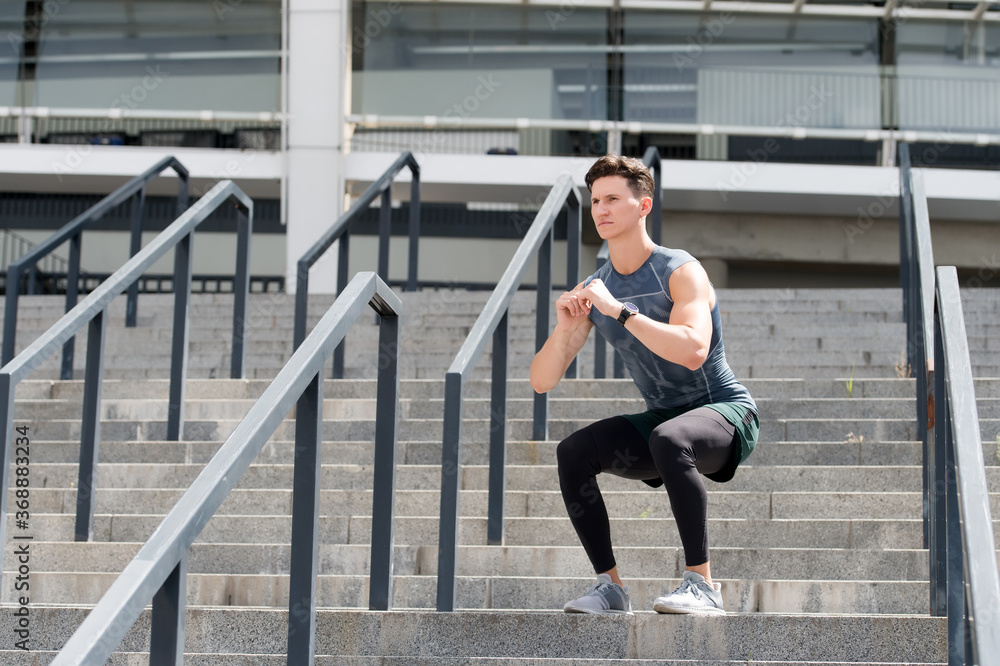 exercising and healthy lifestyle concept. man doing squats outdoor. man with fitness watch exercising and doing squats. Deep squats. young man in sportswear doing squats outdoor