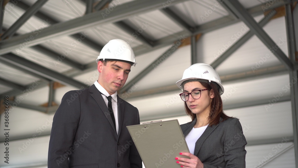 Young man and woman in helmets with documents at a construction site. Chiefs in suits discussing the project of architecture. People look around. Young man and woman in helmets with documents at a construction site. Chiefs in suits discussing the project of architecture. People look around.