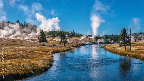 View of the Firehole River flowing through Yellowstone's Upper Geyser Basin, part of the most active geyser field in the world. Old Faithful can be seen erupting in the background.