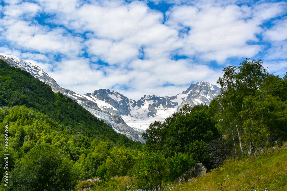 A beautiful view of the snow-capped mountain peaks of the Caucasus Mountains. White clouds against the blue blue sky. A beautiful view of the snow-capped mountain peaks of the Caucasus Mountains. White clouds against the blue blue sky.