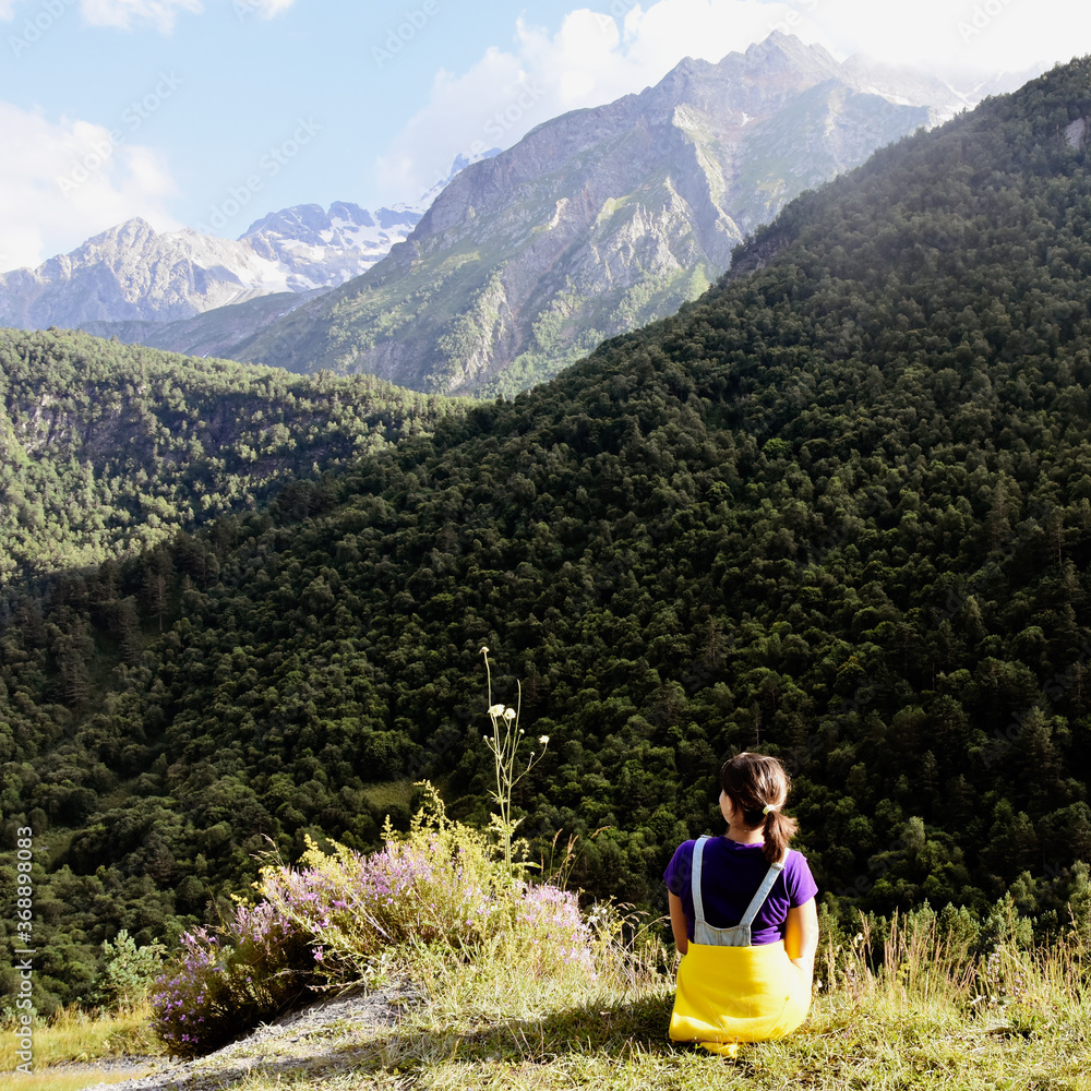 A young girl sits on a cliff and looks at the beautiful snow-capped peaks of the Caucasus Mountains