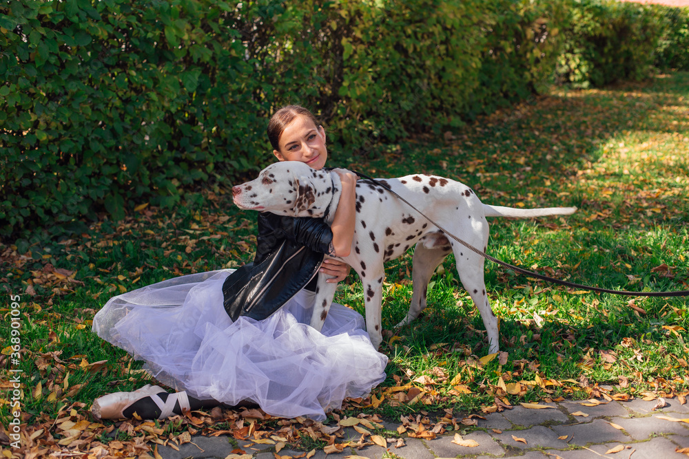 Fototapeta premium Ballerina with Dalmatian dog in the Park. Woman ballerina in a white ballet skirt and black leather jacket and in pointe shoes in autumn park hugging her spotty dalmatian dog.
