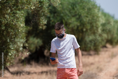 Close up of a young man using a face mask looking down at his phone on an out of focus nature background. Country walk and communication concept.