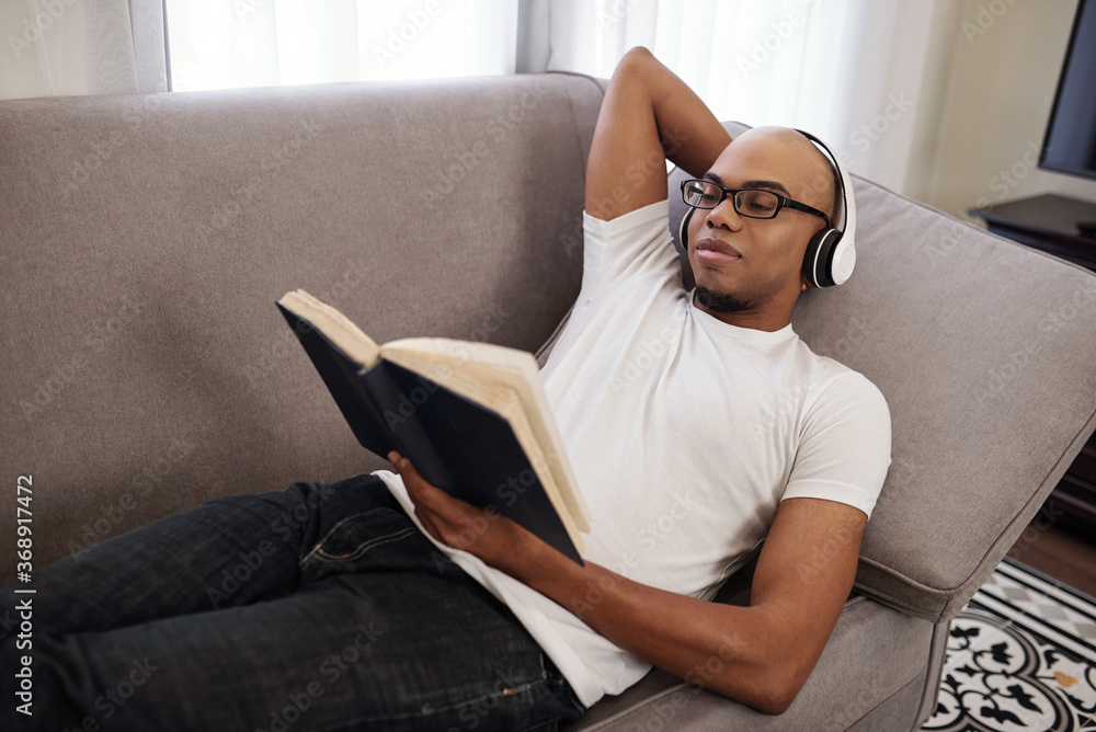 Smiling young African-American man in headphones relaxing on sofa and reading interesting novel Smiling young African-American man in headphones relaxing on sofa and reading interesting novel