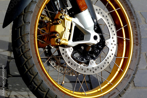 Shiny steel brake double discs on modern large motorcycle wheel. mechanical parts. spokes and brake cable. black rubber tire. transportation safety concept. white tube fork. gray cobblestone pavement