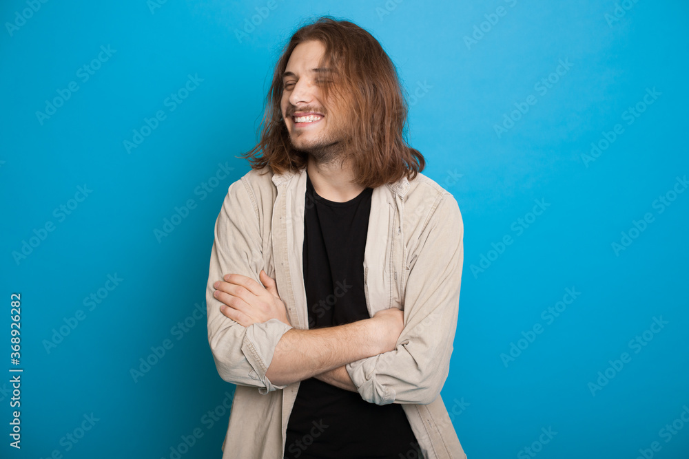 Handsome caucasian man with long hair and beard smiling with crossed hands on a blue studio wall Handsome caucasian man with long hair and beard smiling with crossed hands on a blue studio wall