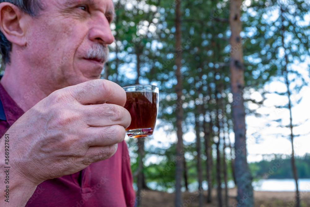 Adult middle-aged man with mustache hold a small glass cup of coffee against the background of the forest or park. Blurred background, selective focus, horizontal orientation. Adult middle-aged man with mustache hold a small glass cup of coffee against the background of the forest or park. Blurred background, selective focus, horizontal orientation.