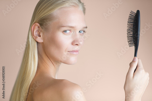 Haircare and hairbrush product advertisement. Beautiful female model with healthy blonde hair holding a brush while looking at the camera. Beauty portrait isolated against a rose gold background.