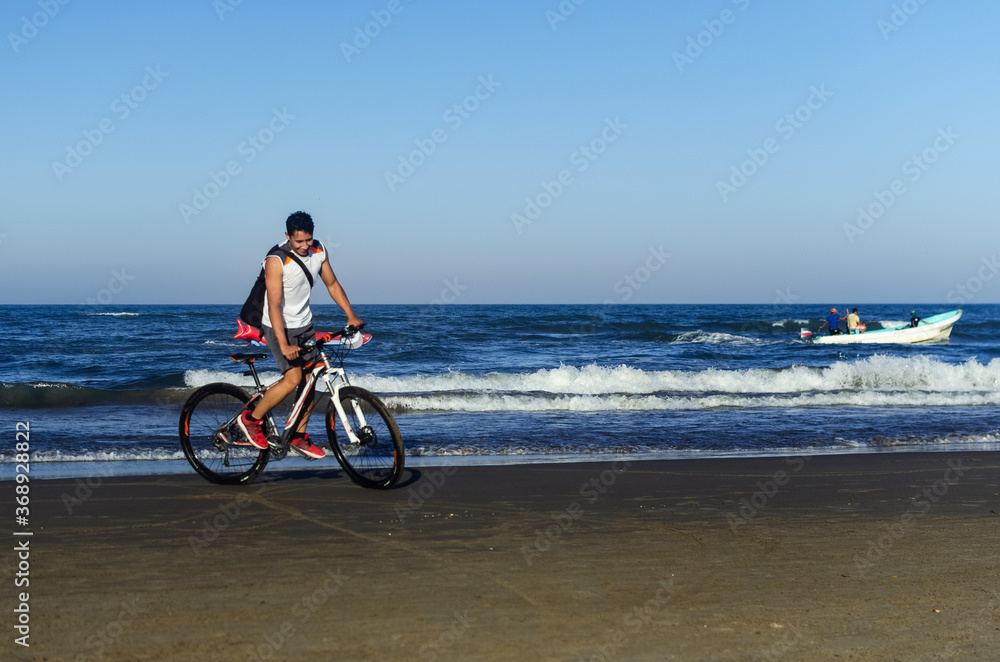 Fototapeta premium joven montado en bici en la playa sonriendo y una lancha de fondo