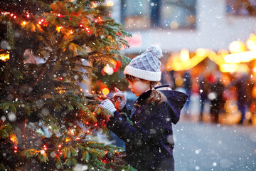Little cute kid girl having fun on traditional Christmas market during strong snowfall. Happy child enjoying traditional family market in Germany. Schoolgirl standing by illuminated xmas tree.