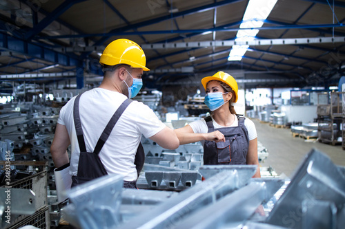 Workers greeting each other with elbow bump due to global corona virus pandemic and danger of infection. Employees standing in factory wearing protection mask.