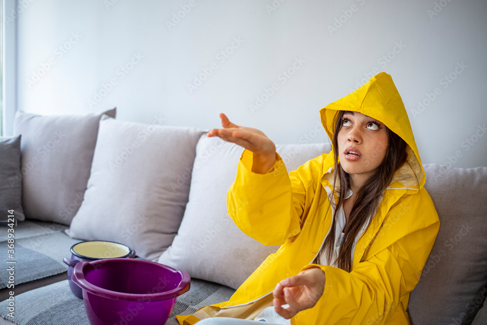 Frustrated homeowner watching ceiling leaks sitting on a couch at home. Time to call plumber. Shocked Woman in raincoat Looks at the Ceiling While Collecting Water Which Leaks Frustrated homeowner watching ceiling leaks sitting on a couch at home. Time to call plumber. Shocked Woman in raincoat Looks at the Ceiling While Collecting Water Which Leaks