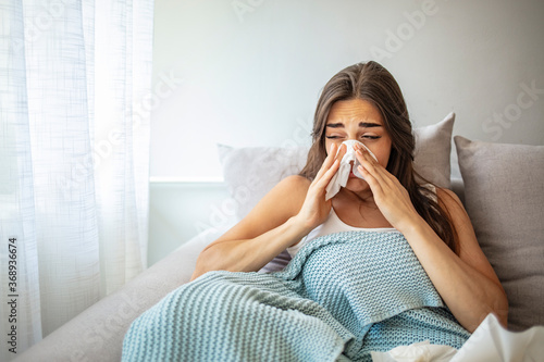 Woman Suffering From Cold Lying In Bed With Tissue. Sick young woman at home on the sofa with a cold, she is covering with a blanket and blowing her nose