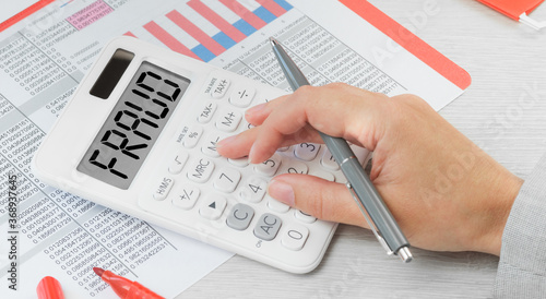 Fraud text on a calculator with financial data paperwork on the desk. Business and finance fraud prevention or identification concept. Hand with calculator