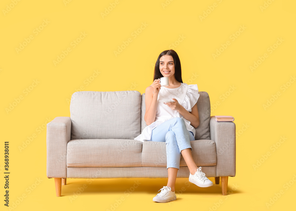 Obraz premium Young woman with tea relaxing on sofa against color background