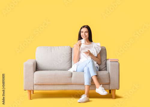 Young woman with tea relaxing on sofa against color background